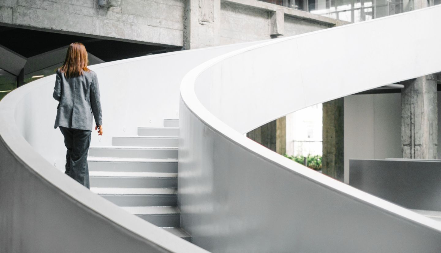 Geschäftsfrau in einem grauen Anzug, die eine weiße, runde Treppe im Bürogebäude hinaufgeht.