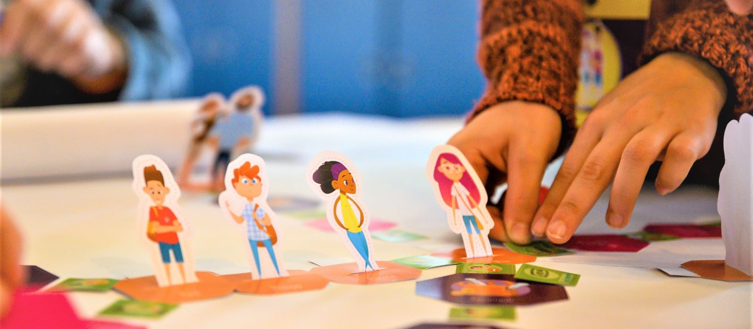Ein Foto zeigt Hände, die Figuren von Schüler*innen auf einem Spielfeld bewegen. Darunter steht der Text: Finanzbildung: Wer früh den richtigen Umgang mit Geld erlernt, ist später besser vor Überschuldung geschützt.