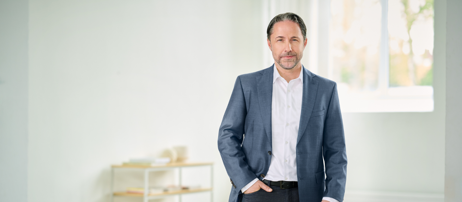 Marwin Ramcke, CEO, steht mit blauem Anzug und der rechten Hand in der Hosentasche in einem hellen Raum. 