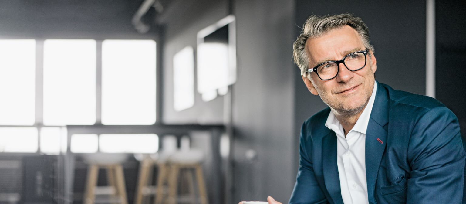 Porträt eines sitzenden, älteren Geschäftsmannes in einem blauen Anzug, der eine weiße Kaffeetasse hält.