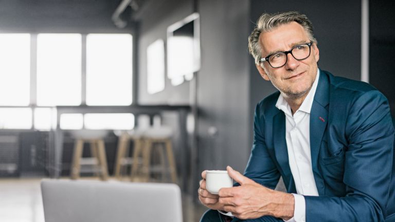 Porträt eines sitzenden, älteren Geschäftsmannes in einem blauen Anzug, der eine weiße Kaffeetasse hält.
